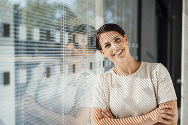 Smiling businesswoman with arms crossed leaning on window - JOSEF20728