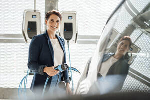 Lächelnde Geschäftsfrau mit Elektrostecker am Auto an der Ladestation - JOSEF20765