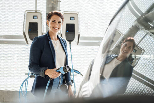 Smiling businesswoman with electric plug by car at charging station - JOSEF20765