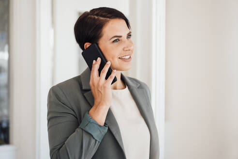 Lächelnde Geschäftsfrau, die im Büro mit einem Smartphone telefoniert - JOSEF20793