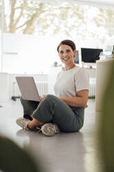 Happy businesswoman with laptop sitting on floor at office - JOSEF20814