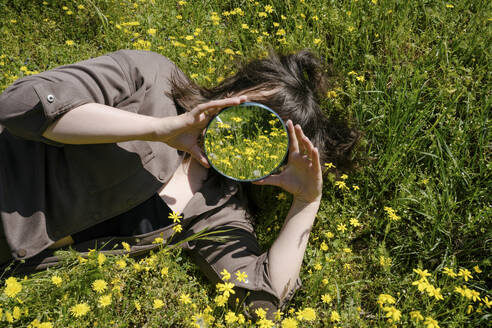 Frau hält Spiegel über Gesicht und liegt im Gras auf einem Feld - YBF00192