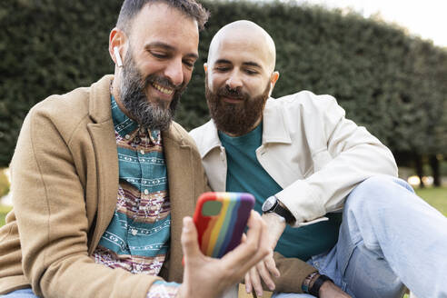 Happy gay man using smart phone with friend in park - JPTF01310