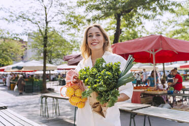 Happy woman standing with fresh leafy vegetables and lemons in market - NDEF01001