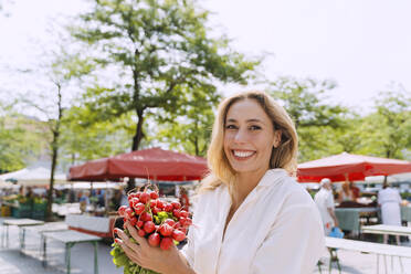 Glückliche blonde Frau hält Rettich auf dem Bauernmarkt - NDEF01005