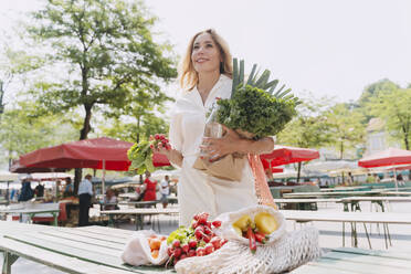 Smiling woman with various vegetables standing at table in market - NDEF01010