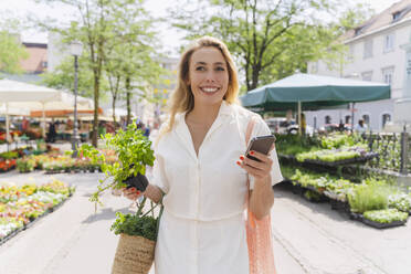 Glückliche Frau hält Minzpflanze und Smartphone an einem sonnigen Tag - NDEF01042
