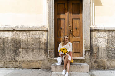 Lächelnde Frau sitzt auf Stufen mit einem Strauß Sonnenblumen vor einer Holztür - NDEF01058