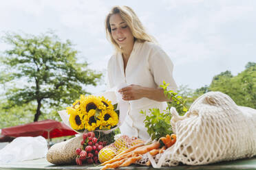 Happy woman with bunch of sunflowers and groceries at table in market - NDEF01068