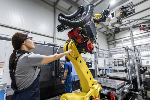 Technician controlling robotic arm with colleague operating machinery in background - AAZF00939