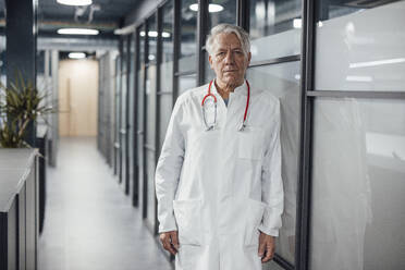 Senior doctor with stethoscope leaning on glass wall in clinic - JOSEF20931