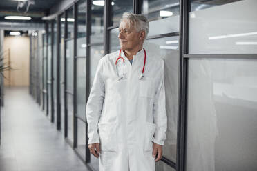 Smiling doctor with lab coat leaning on glass wall at hospital - JOSEF20932