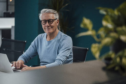 Lächelnder älterer Geschäftsmann mit Brille arbeitet am Schreibtisch im Büro - JOSEF20952