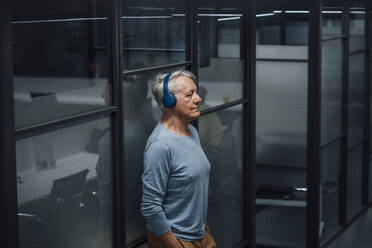Senior businessman listening music on headphones at work place - JOSEF20955