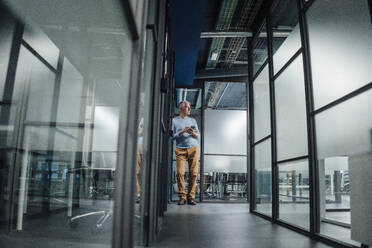 Thoughtful businessman holding smart phone in office corridor - JOSEF20959