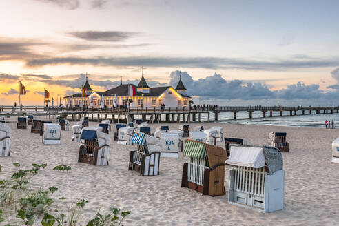 Germany, Mecklenburg-Vorpommern, Ahlbeck, Hooded beach chairs at sunset with pier and bathhouse in background - EGBF00897