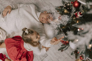 Verspielter Vater und Tochter liegen auf dem Boden und berühren den Weihnachtsbaum - VIVF01068