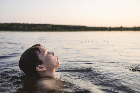 Junge spuckt Wasser in See bei Sonnenuntergang - EYAF02798