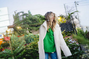 Blond woman carrying Christmas tree at market at dusk - MDOF01470