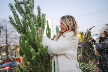 Smiling woman touching Christmas tree at market - MDOF01476