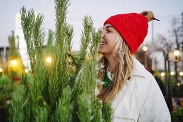 Lächelnde Frau mit Strickmütze in der Nähe des Weihnachtsbaums auf dem Markt - MDOF01480