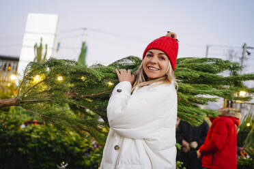 Smiling woman wearing knit hat carrying Christmas tree at market - MDOF01481