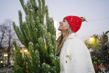 Smiling woman wearing knit hat standing next to Christmas tree at market - MDOF01483