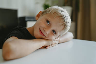 Lächelnder Junge, der sich am Tisch im Haus ausruht - ANAF02103