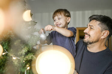 Smiling father and son decorating Christmas tree at home - ANAF02111