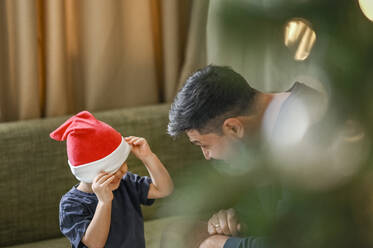 Playful son covering face with santa hat on sofa at home - ANAF02116