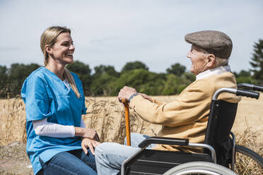 Smiling nurse talking with elderly man sitting in wheelchair on sunny day - UUF30198