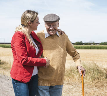 Happy elderly man walking with woman by field on sunny day - UUF30206