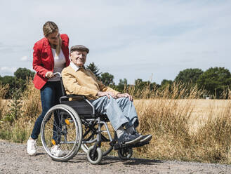 Woman pushing elderly man sitting in wheelchair by field - UUF30211