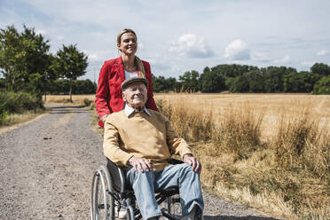 Frau schiebt einen älteren Mann im Rollstuhl durch ein Feld - UUF30213