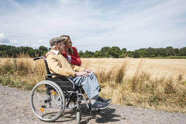 Älterer Mann sitzt im Rollstuhl neben einer Frau und schaut auf ein Feld - UUF30217