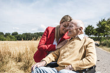 Glückliche Frau mit älterem Mann im Rollstuhl sitzend an einem sonnigen Tag - UUF30218