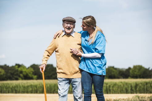 Senior man laughing and taking support of healthcare worker on sunny day - UUF30227