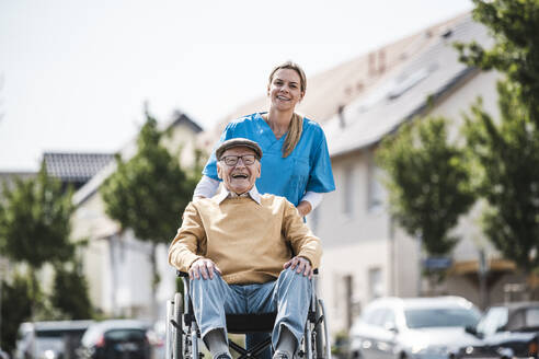 Cheerful senior man sitting in wheelchair and nurse pushing him on sunny day - UUF30230