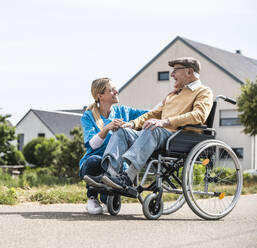 Lächelnde Krankenschwester im Gespräch mit einem älteren Mann im Rollstuhl an einem sonnigen Tag - UUF30235