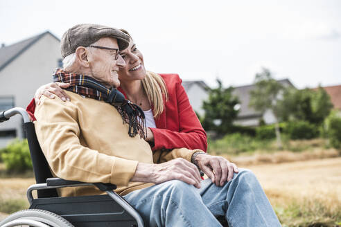 Lächelnde Frau umarmt älteren Mann, der im Rollstuhl sitzt - UUF30247