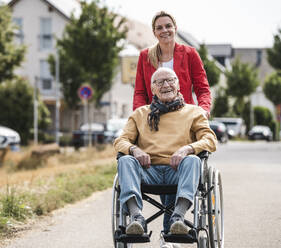 Smiling woman pushing senior man sitting in wheelchair on road - UUF30253