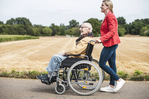 Frau schiebt glücklichen älteren Mann im Rollstuhl sitzend auf der Straße - UUF30258