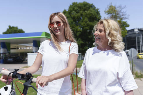 Glückliche ältere Frau mit Tochter, die an einem sonnigen Tag einen Elektroroller hält - AAZF00996