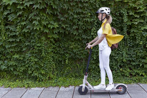 Junge Frau fährt mit einem Elektroroller vor Pflanzen - AAZF01007