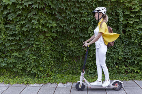 Young woman riding electric push scooter in front of plants - AAZF01007