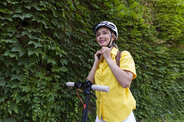 Lächelnde junge Frau, die ihren Helm einstellt und vor Pflanzen steht - AAZF01012