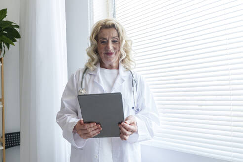 Lächelnder Oberarzt mit Tablet-PC am Fenster in einer Klinik - AAZF01027