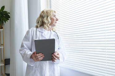 Thoughtful senior doctor with tablet PC looking through window - AAZF01028
