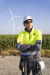 Contemplative engineer standing with arms crossed in front of wind turbines - EKGF00505