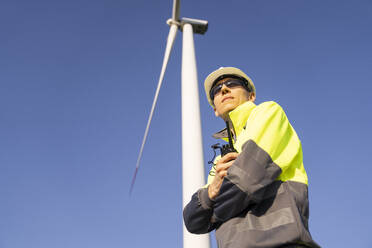 Thoughtful engineer standing with arms crossed under wind turbine - EKGF00507